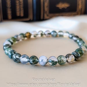May include: A beaded bracelet with translucent, round beads featuring green and white inclusions. The bracelet is displayed on a cream-coloured surface, with a blurred background of dark books. The beads are approximately 0.8 cm in diameter.