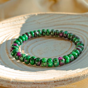 Peut inclure: Un bracelet de perles composé de pierres précieuses vertes et magenta. Le bracelet est fait de petites perles rondes enfilées ensemble. Les perles ont un aspect moucheté, avec un mélange de couleurs vertes et magenta. Le bracelet est présenté sur une surface en bois.