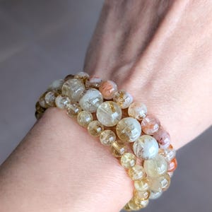 Op de afbeelding: Drie armbanden met heldere, gele en oranje gekleurde edelstenen. De armbanden zijn op een pols gestapeld, waarbij de verschillende maten van de kralen te zien zijn. De kralen zijn doorschijnend, wat een natuurlijke en aardse uitstraling geeft.