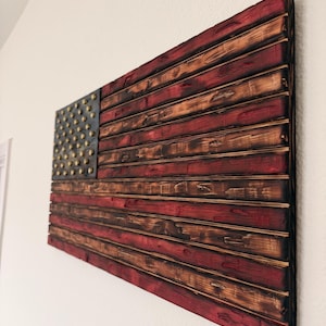 May include: A rectangular wooden wall art piece designed as a flag with horizontal slats. The surface features a distressed, burnt finish with alternating red and natural wood tones. A dark blue canton section displays raised metallic studs.