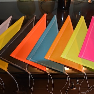 May include: Nine triangular fabric flags in various colors, including white, yellow, black, orange, teal, yellow, green, pink, and neon pink. The flags are arranged in a row, with the white flag at the left and the neon pink flag at the right. Each flag has a white stitched border.