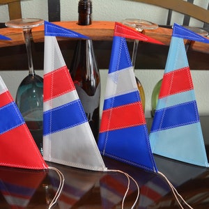 May include: Four triangular fabric sailboat ornaments featuring stitched panels in red, white, and blue color combinations. Each ornament has a small triangular flag at the top and a thin string attached to the bottom corner for hanging.