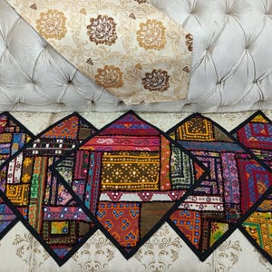 May include: A cream-colored tufted sofa with a decorative floral patterned runner and five colorful patchwork cushions. The cushions have a black border and feature a variety of patterns and colors, including red, purple, and gold.