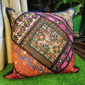 May include: Square cushion with a patchwork design in shades of pink, orange, and purple. The cushion has embroidered details and small mirrored accents. It is placed on a green surface. The cushion is a decorative item.