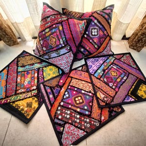 May include: A selection of colourful, patchwork-style cushions and square placemats. The textiles feature vibrant hues of pink, purple, orange, and yellow, with intricate patterns and a black border. The cushions are triangular, and the placemats are square.