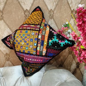 May include: A square decorative pillow with a patchwork design. The pillow features a variety of colors, including yellow, black, red, blue, and orange. The patchwork includes embroidered details and small mirrored accents. The pillow is placed on a white sofa.