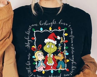 May include: Black t-shirt featuring a Christmas-themed graphic with the Grinch and other characters. The design includes a frame of Christmas lights and text that reads "Little bit more, Maybe christmas means a perhaps. I come from a store maybe christ was, hethought. doesn't".