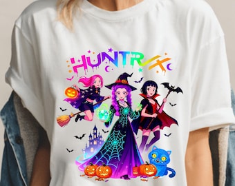 May include: White t-shirt featuring a colorful graphic of three cartoon witches with pumpkins, bats, and a blue cat. The word "HUNTRX" is printed above the witches in a rainbow gradient.