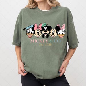 May include: An olive green t-shirt featuring the faces of Disney characters Donald Duck, Minnie Mouse, Mickey Mouse, and Daisy Duck. The text "MICKEY & CO Est. 1928" is printed below the characters in pastel colors. The shirt has short sleeves.