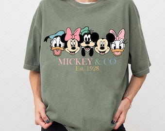 May include: An olive green t-shirt featuring the faces of Disney characters Donald Duck, Minnie Mouse, Mickey Mouse, and Daisy Duck. The text "MICKEY & CO Est. 1928" is printed below the characters in pastel colors. The shirt has short sleeves.