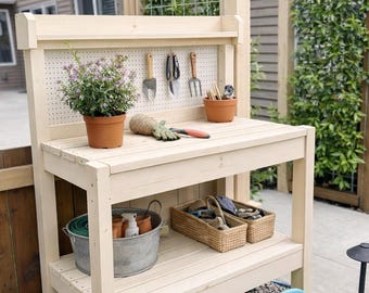 May include: A light-colored wooden potting bench with a pegboard back, holding gardening tools and a potted plant. The bench has a lower shelf with a galvanized bucket, baskets, and a blue watering can nearby. The structure is outdoors.