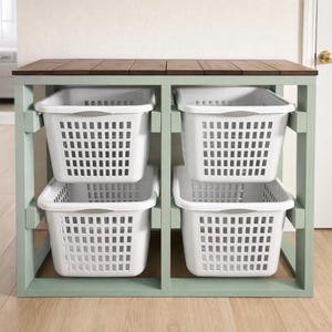 May include: A four-compartment storage unit with four white plastic laundry baskets. The unit has a light green frame and a dark brown wooden top. The baskets are rectangular with a grid pattern and gray handles.