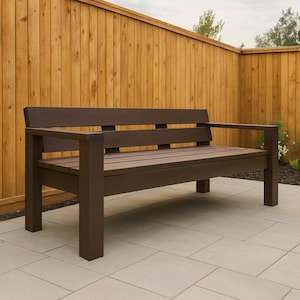 以下が含まれることがあります： アームレストとスラットシートが付いた茶色の木製ベンチ。ベンチは屋外にあり、木製のフェンスとタイルのパティオを背景にしています。ベンチは濃い茶色です。