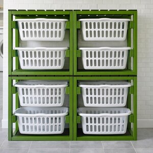 May include: A green wooden laundry sorter with six white plastic laundry baskets. The sorter is in a laundry room setting with a washing machine and dryer in the background. The baskets are rectangular with slotted sides.