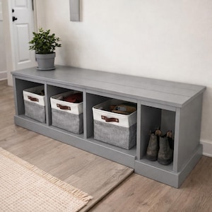 Puede incluir: Un banco de almacenamiento de madera gris con tres compartimentos. Tres cestas de almacenamiento de tela gris y blanca con asas de cuero marrón están en los tres primeros compartimentos. Una pequeña planta en maceta está encima. Botas en el último compartimento.