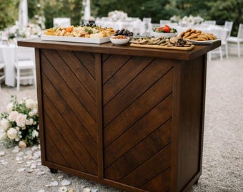 May include: A dark brown wooden bar with a chevron pattern on the front. The bar is on wheels and has a countertop with various food items, including cheese, grapes, and crackers. The bar is set up outdoors, likely for an event.