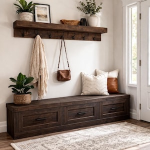 Peut inclure: Un banc d'entrée en bois foncé avec trois tiroirs, surmonté de coussins décoratifs. Au-dessus, une étagère avec des crochets supporte un sac à main et une couverture. Des plantes et un tableau encadré complètent le décor.