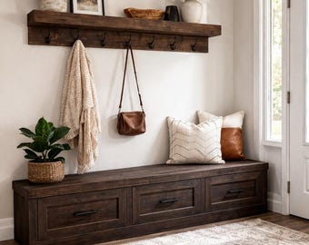 May include: A dark wood entryway bench with three drawers, topped with decorative pillows. Above, a shelf with hooks holds a purse and a blanket. Plants and a framed picture add to the decor. A rug is on the floor.