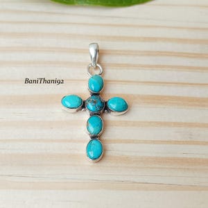 May include: A silver-toned pendant in the shape of a cross, adorned with oval and round turquoise stones. The pendant has a bail at the top for a chain. The background is a light-colored wooden surface. The text "BaniThani92" is visible.