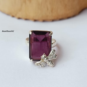 Könnte beinhalten: Ein Silberring mit einem großen, rechteckigen, tiefvioletten Edelstein. Ein detaillierter silberner Bienenanhänger ist am Ringband befestigt. Der Ring ist vor einem weißen Hintergrund mit einem Holzelement im Hintergrund platziert.