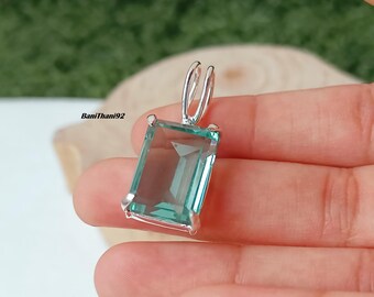 May include: A rectangular, aquamarine-colored pendant with a silver-colored bail. The gemstone is set in a silver-colored metal frame. The pendant is held in a hand against a blurred background.