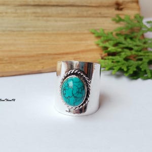 May include: A wide silver ring with a large oval turquoise stone. The stone is set in a twisted rope-like border. The ring has a polished finish and is displayed on a white surface.