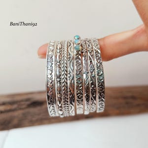 May include: A stack of silver bangles with intricate floral and leaf patterns. Some bangles have small, light blue gemstones. The bangles are held by a person's finger. The text "BaniThani92" is in the upper left corner.