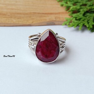May include: A silver ring featuring a large, teardrop-shaped, deep red gemstone. The ring has a multi-band design on the band. The gemstone is the focal point, showcasing its rich colour and polished surface. The ring is set against a neutral background.
