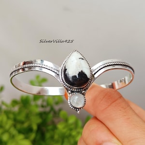 May include: A silver-toned bracelet featuring a teardrop-shaped white and black stone as the centerpiece, with a smaller, round, light-colored stone below. The bracelet has a unique design with open bands on each side. The text "SilverVilla925" is visible.