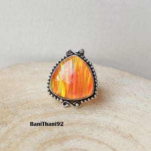 May include: A silver ring with a large, triangular, orange and yellow gemstone. The gemstone has a vibrant, striped pattern. The ring has a detailed, beaded border and decorative accents. The text "BaniThani92" is at the bottom.