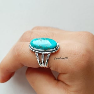 May include: A silver ring featuring a large, oval turquoise stone. The stone is a vibrant blue-green color, set in a detailed silver band with a braided edge. The ring has a split shank design.