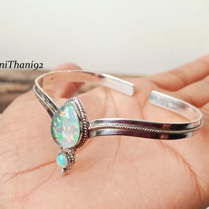 Puede incluir: Un brazalete plateado con una piedra de ópalo en forma de pera y un ópalo redondo más pequeño. El brazalete tiene un diseño delicado en forma de cuerda a lo largo de la banda y está abierto en la parte posterior. El texto "BaniThani92" es visible.