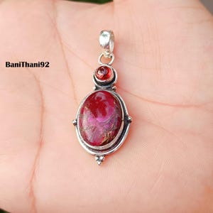 Pode incluir: Pingente de prata com uma grande pedra preciosa vermelha oval e uma pedra preciosa vermelha redonda menor. O pingente tem uma moldura decorativa de prata e uma argola para uma corrente. O texto "BaniThani92" é visível no lado esquerdo da imagem.