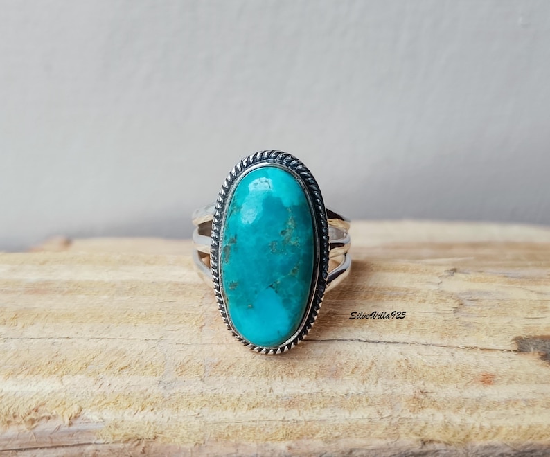 May include: A silver ring with a large, oval turquoise stone. The stone is a vibrant blue-green color with natural variations. The ring has a braided border around the stone and a triple-band design. The ring is displayed on a wooden surface.