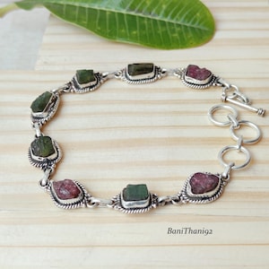 Peut inclure: Un bracelet en argent orné de pierres précieuses brutes vertes et roses alternées. Le bracelet est doté d'un fermoir à bascule et est présenté sur une surface en bois clair. Les pierres sont serties dans des lunettes en argent ornées.