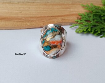 May include: A silver ring with an oval-shaped stone. The stone features a mosaic of colors, including turquoise, orange, white, and gold. The ring has a wide band and a textured surface. The ring is displayed on a white surface with a wooden background and green foliage.