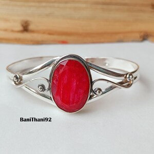 Puede incluir: Brazalete plateado con una gran piedra preciosa roja brillante de forma ovalada en el centro. El brazalete tiene un diseño calado con detalles en espiral y pequeños acentos redondos. El texto "BaniThani92" es visible.