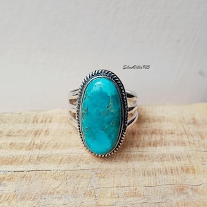 May include: A silver ring featuring a large, oval turquoise stone. The stone is a vibrant shade of blue-green with natural matrix patterns. The ring has a braided border around the stone and multiple bands on the sides. The ring is displayed on a wooden surface.