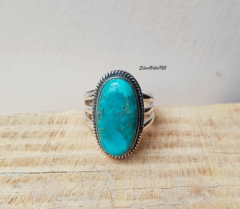 May include: A silver ring featuring a large, oval turquoise stone. The stone is a vibrant shade of blue-green with natural matrix patterns. The ring has a braided border around the stone and multiple bands on the sides. The ring is displayed on a wooden surface.