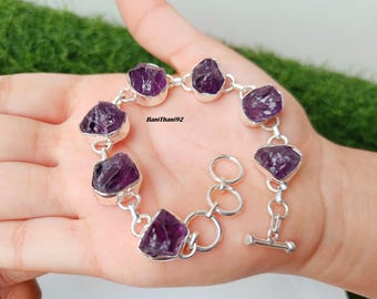 May include: A silver bracelet featuring raw, deep purple amethyst stones. The stones are set in individual silver bezels and connected by silver links. The bracelet is displayed on a hand against a green background.