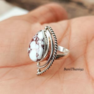 May include: A sterling silver ring featuring a teardrop-shaped white stone with maroon veining. The stone is set in a decorative silver frame with a twisted rope design along the side. The ring is a unique piece of jewelry.