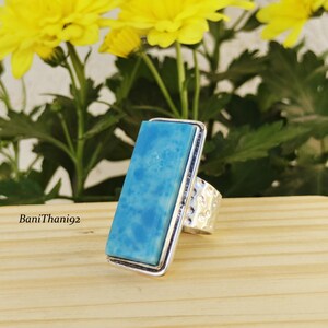May include: A rectangular ring with a light blue stone set in a silver frame. The ring has a textured band and is displayed on a wooden surface with yellow flowers in the background. The text "BaniThani92" is visible.