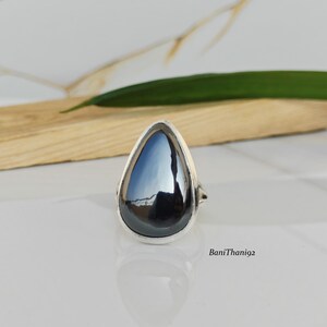 Puede incluir: Un anillo de plata con una gran piedra preciosa negra pulida en forma de lágrima. El engaste del anillo es de plata brillante, y la piedra preciosa refleja la luz. El anillo se exhibe sobre una superficie blanca con una tabla de madera y una hoja verde en el fondo.