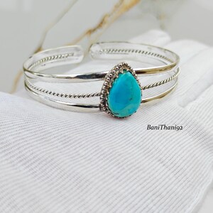 May include: A silver cuff bracelet featuring a central teardrop-shaped turquoise stone set in a beaded bezel. The band consists of three parallel silver wires, with the outer two featuring a twisted rope texture and the centre being smooth.