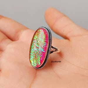 Op de afbeelding: Een zilveren ring met een ovale, iriserende steen. De steen toont een levendige mix van roze, groen en geel. De ring heeft een getextureerde, touwachtige rand en een gespleten schachtontwerp.