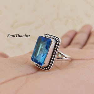 May include: A rectangular blue gemstone ring with a silver band. The gemstone is set in a silver frame with a beaded border. The ring is displayed on a hand.