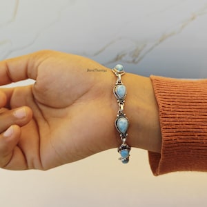 May include: A silver bracelet with five light blue, teardrop-shaped stones. The bracelet is worn on a wrist with an orange long-sleeved shirt. The stones are set in ornate silver settings, and the bracelet is connected by silver links.