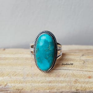 May include: A silver ring with a large, oval turquoise stone. The stone is a vibrant blue-green color and is set in a silver band with a rope-like border. The ring is on a wooden surface.