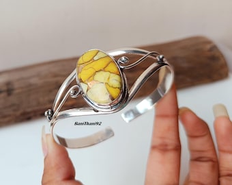 May include: A silver cuff bracelet featuring an oval-shaped, yellow and brown stone. The stone is the focal point, set in a decorative silver frame. The bracelet has an open design, allowing for easy wear.