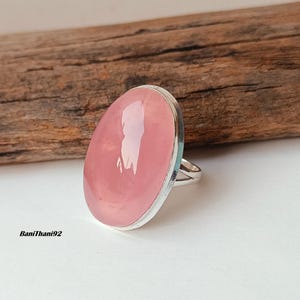 May include: A silver ring featuring a large, oval rose quartz gemstone. The pink stone is set in a silver bezel. The ring is displayed on a white surface with a piece of wood in the background. The text "BaniThani92" is visible.